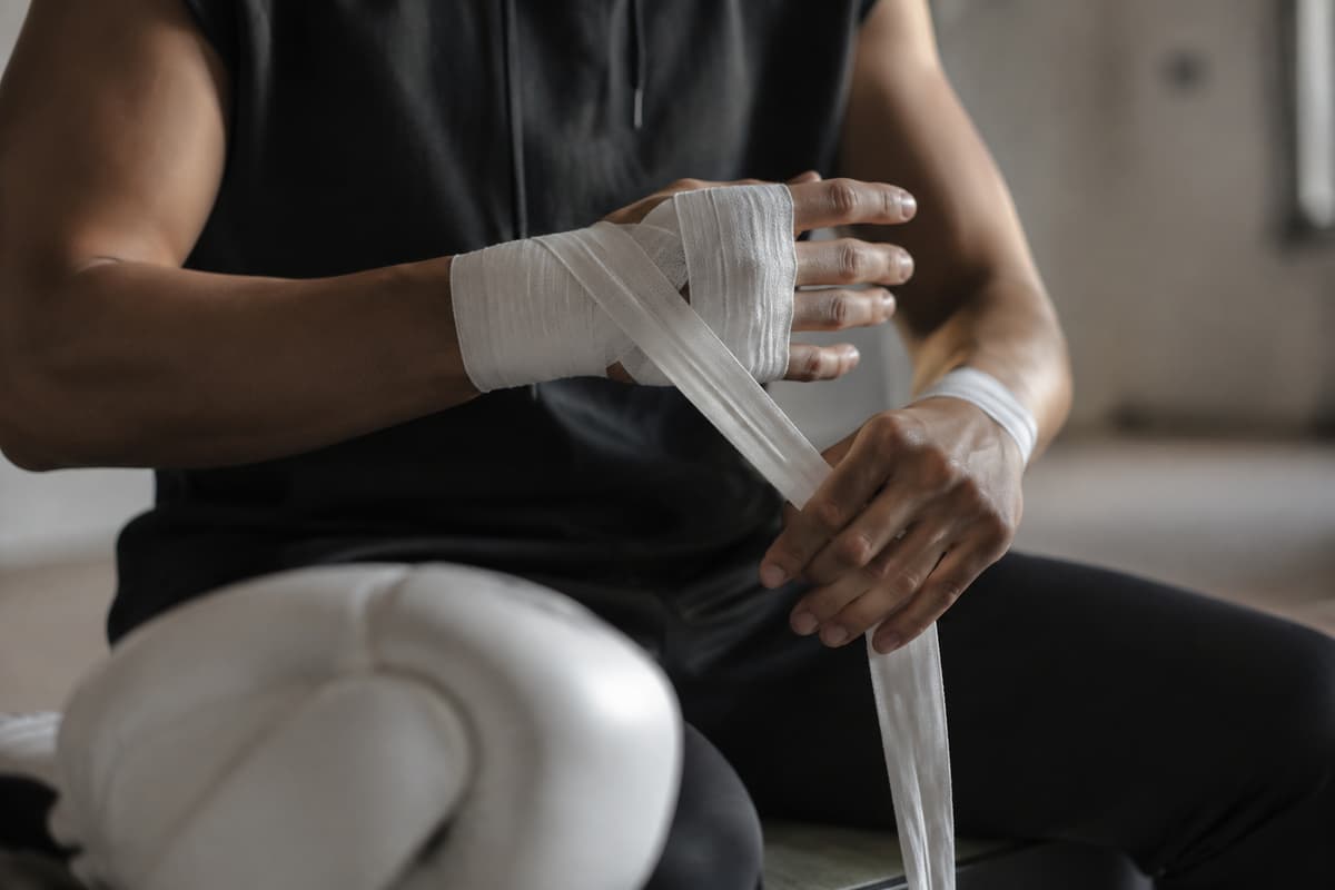 Un boxeur adulte ajuste une bande de boxe propre autour de son poignet, avec une main partiellement bandée et un gant de boxe posé au premier plan.